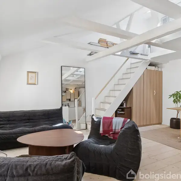 Stue med sorte loungestole og rundt sofabord. En hvid trappe fører til en hems. Der er et spisebord med tre stole og en plante i hjørnet.
