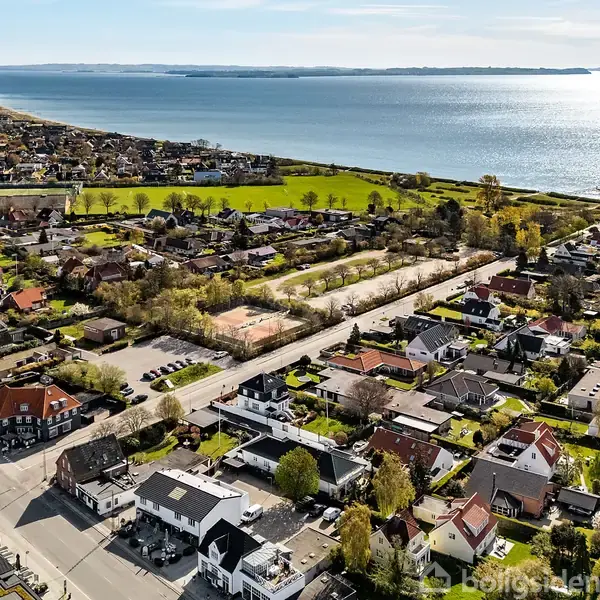 Fugleperspektiv af et boligområde langs kysten. Huse og træer ligger tæt ved vejen, mens havet strækker sig ud i baggrunden, hvilket skaber en naturskøn kystnær udsigt.