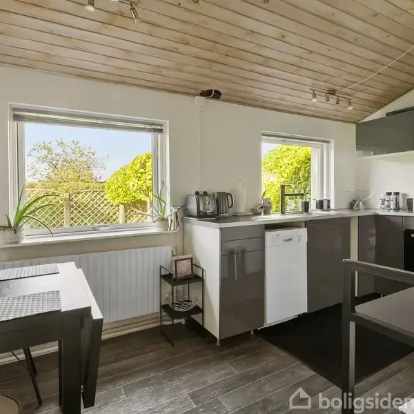 Køkken med grå skabe og hvide vægge indeholder komfur, opvaskemaskine og køkkenudstyr. To vinduer giver udsigt til grønne buske udenfor. Et lille spisebord står i rummet.