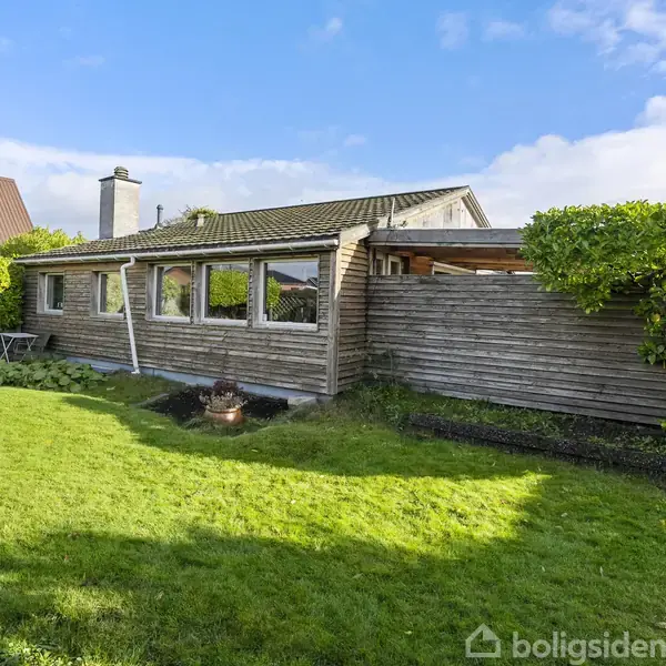 En lav, lang træbygning med flere vinduer står stille i en grøn have med velplejede buske og græsplæne. Blå himmel og nabohuse skaber en rolig nabolagsstemning.