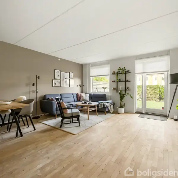 Stue med grå sofa og stol placeret omkring et sofabord på et tæppe. Vægdekorationer hænger over sofaen, og der er reoler med planter ved en glasskydedør til en have.