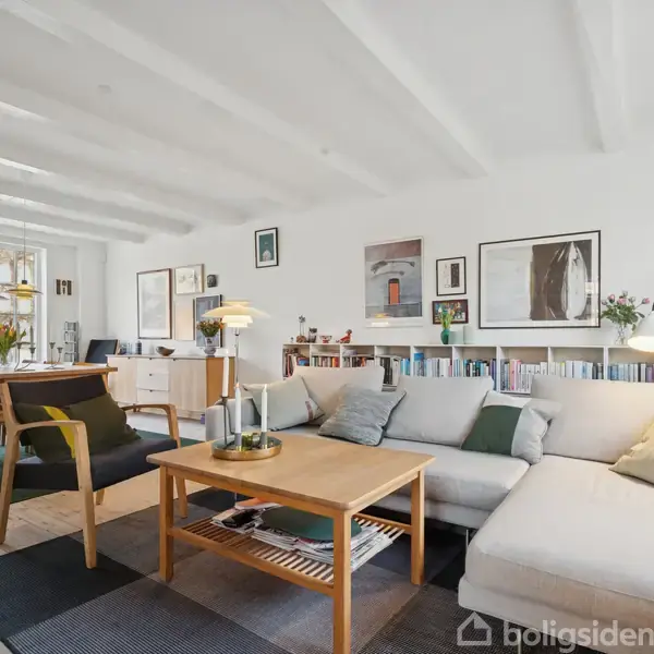 Stor, lys stue med lyse træmøbler og grå hjørnesofa. Aktion: Rolig og indbydende atmosfære. Kontekst: Vægdekorationer, bøger, lamper og planter skaber hygge. Ingen tekst i billedet.