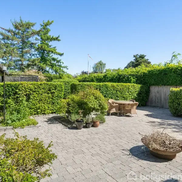 En terrasse med fliser og planter i krukker. Midt i terrassen står et ildsted. Til højre ses et rundt bord med stole. Grønt buskads omkranser området.