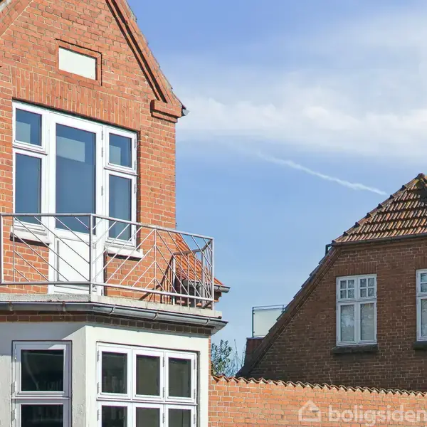 Rødt murstenshus med en altan foran to vinduer, set i en beboelseskontekst med en anden bygning i baggrunden under en blå himmel.