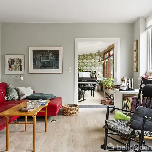 Rød sofa står mod væggen, suppleret af et træbord foran den i en stue med kunst på væggene. I baggrunden ses et rum med klaver og grøn tapet.