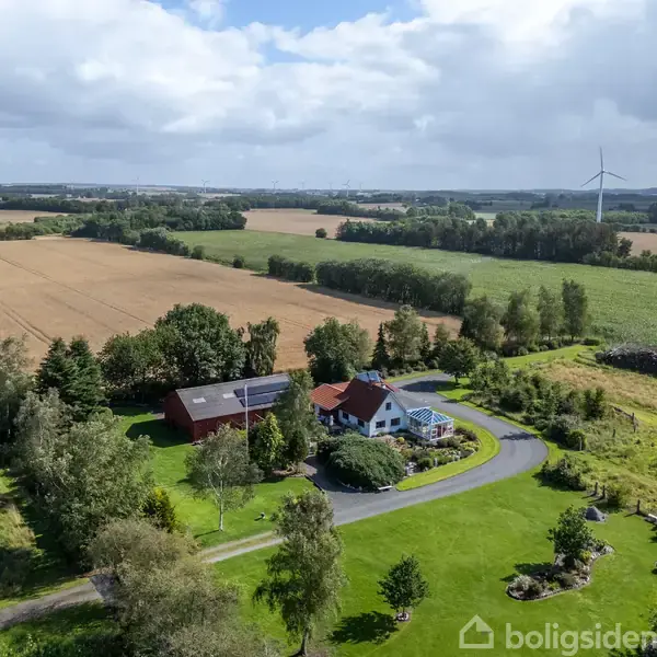 Et hus med rødbrunt tag omgivet af grønne træer og marker. En vindmølle ses i baggrunden under en skyet himmel. Tekst: "boligsiden" er i hjørnet.