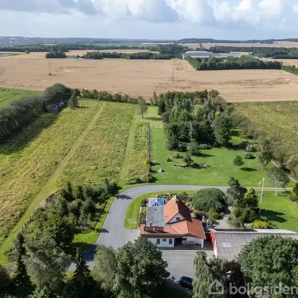 Et hus med rødtag ligger i det åbne landskab omgivet af frodig have. Omkring huset ses store marker og træer under en blå himmel med skyer.