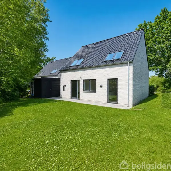 En moderne hvid murstensvilla står stille på en velholdt grøn græsplæne. Store vinduer og et sort skråt tag pryder huset. Træer omgiver ejendommen under en klar blå himmel.