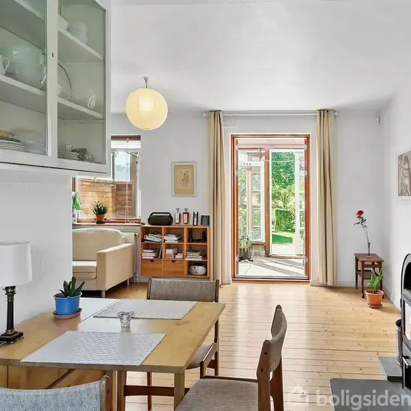 Et spisebord med fire stole står i et rum med lyse trægulve. En brændeovn er til højre, og glasskydedøre fører til en terrasse med udsigt til haven.