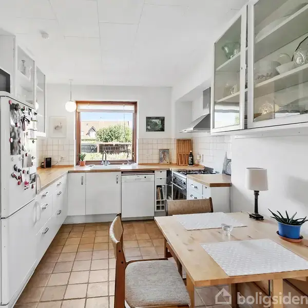 Køkkenbord med to stole står foran et stort vindue, omgivet af hvide skabe og apparater på gulv med brune fliser. Kaktus og bordlampe på bordet.