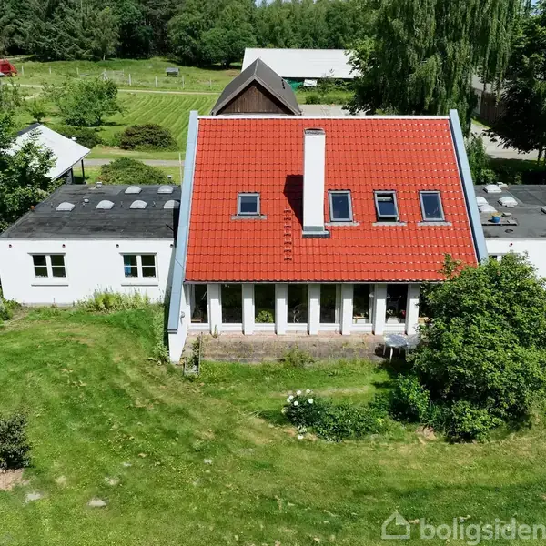 Et hus med rødt tag og hvid facade står omgivet af grønne græsplæner og træer, langt fra andre bygninger i en landlig, skovrig omgivelse.