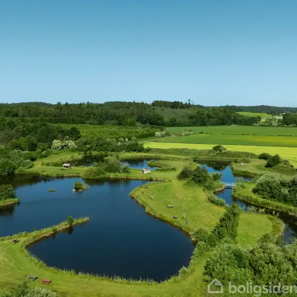 Søer omkranset af grøn vegetation ligger stille i et åbent landskab af marker og skove under en klar blå himmel.