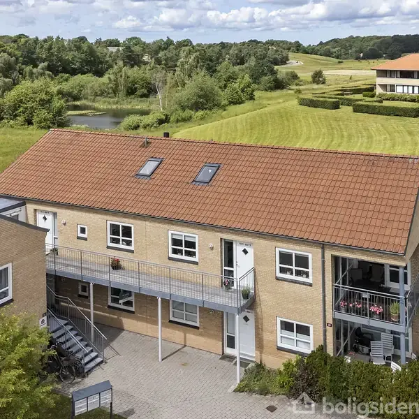 En murstensbygning med rødt tegltag står i en grøn, landlig omgivelser. Bygningen har udvendige trapper og balkon. Baggrunden viser en mark og skovområder med en lys daghimmel.