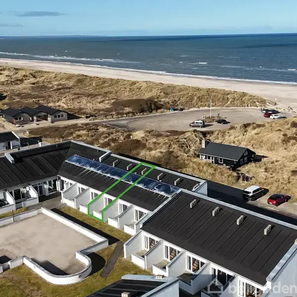 Boligkompleks med sorte tage strækker sig langs klitterne ved havet. Solpaneler er installeret på taget. Omkringliggende natur inkluderer strand og spredte huse. Skyfri, blå himmel mod horisonten.