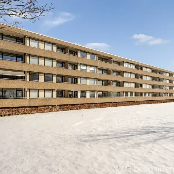 En lang, rektangulær boligblok står stille ved en snedækket flade, omgivet af nogle spredte træer og en blå himmel.