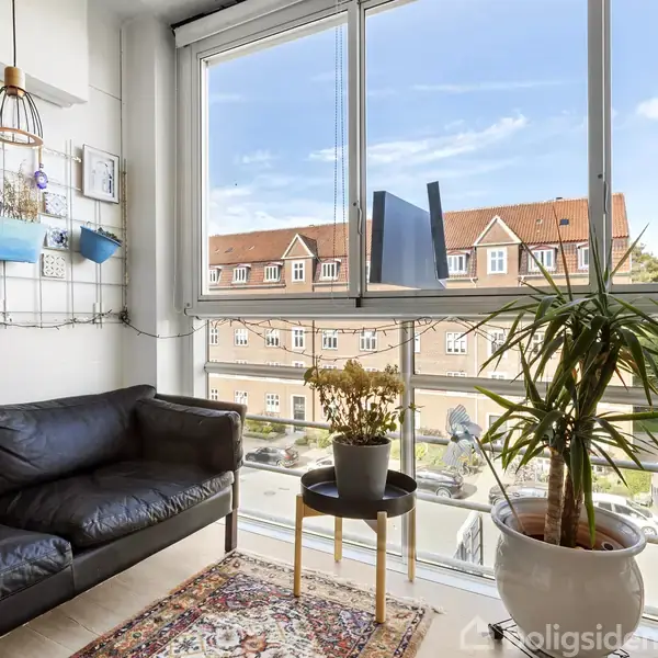 Sort sofa står ved glasvinduer i en lys stue, med en udsigt over røde tage. To planter er placeret ved siden af et lille bord på et tæppe.