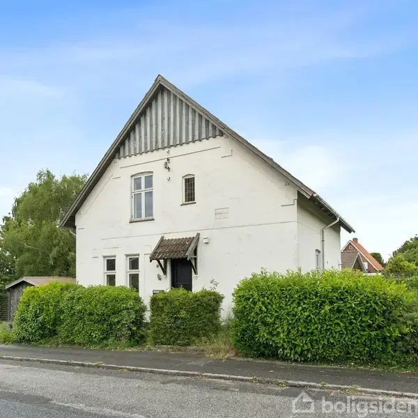 Hvidt hus med gavl og skråt tag står stille ved vejen, omkranset af grønne buske og træer under en klar, blå himmel.