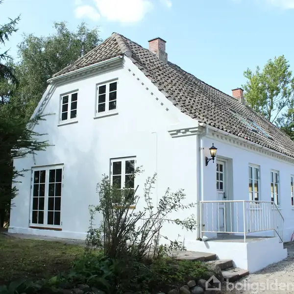 Hvidt hus med døre og vinduer, står i en have omgivet af træer. Trin fører op til hoveddøren, og en lanterne hænger ved indgangen.
