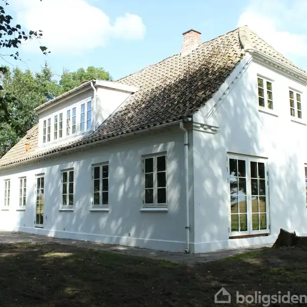 Et hvidt hus med skråt tag står stille i sollys. Det er omgivet af træer og en græsplæne, hvilket skaber en fredelig atmosfære.