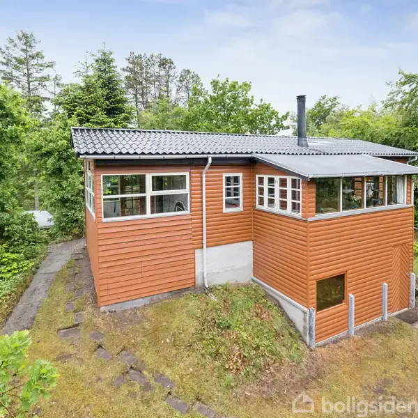 Et træhus med rødbrune paneler står på en græsklædt bakke omgivet af tæt skov. Huset har store vinduer og et sort tag med en skorsten.