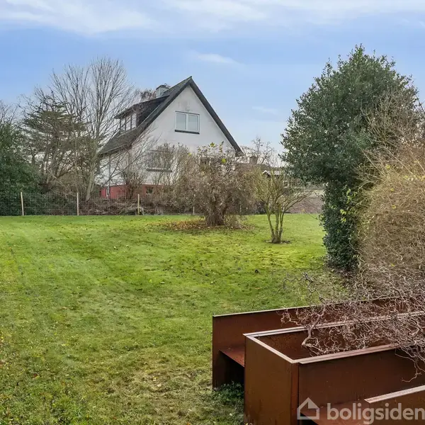 Et hus med spidst tag står i en have omgivet af buske og træer. Foran er der en græsklædt have med visne buske og en metalstruktur til højre.