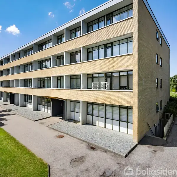 En modernistisk, firkantet bygning med fire etager og store vinduespartier står i et grønt område under en klar blå himmel. En sti fører op til bygningens indgang.