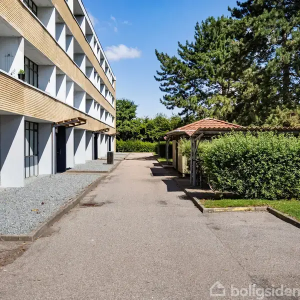 En moderne lejlighedsbygning med store vinduer strækker sig langs en asfalteret sti. Til højre ses en lille have med buske og et halvtag.