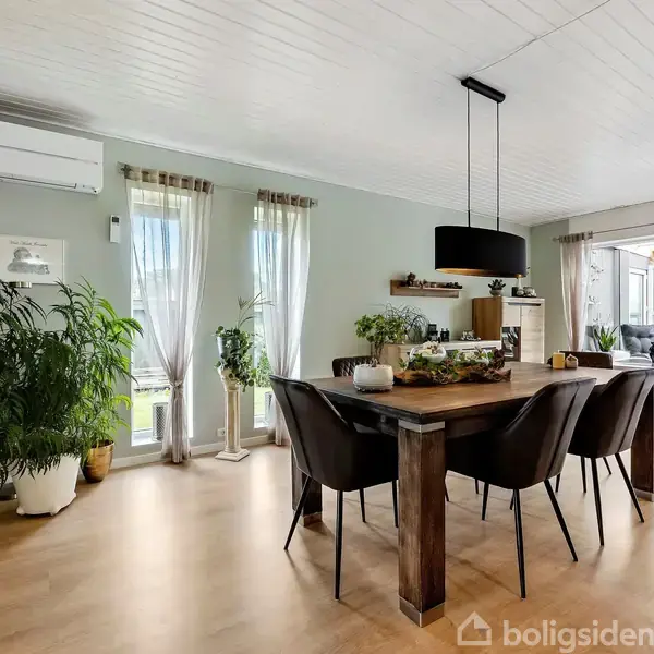Spisebord med mørke stole står i et lyst rum med trægulv. Store planter og vinduer med lette gardiner omkranser. Sort pendel hænger over bordet. Udenfor ses en veranda.