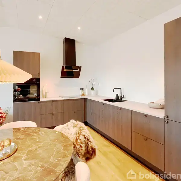 Køkken med træskabe står stille i et moderne hjem. Omkring er der en rund bordplade med dekorationer, lamper hænger fra loftet, og væggene er hvide.