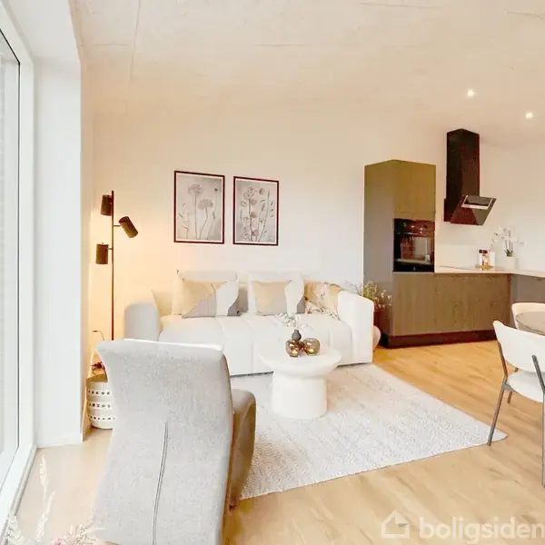 Stue med en sofa, to billeder på væggen, ved siden af et moderne køkkenhjørne med spisebord og fire stole. Rummet har trægulv og et stort vindue til venstre.