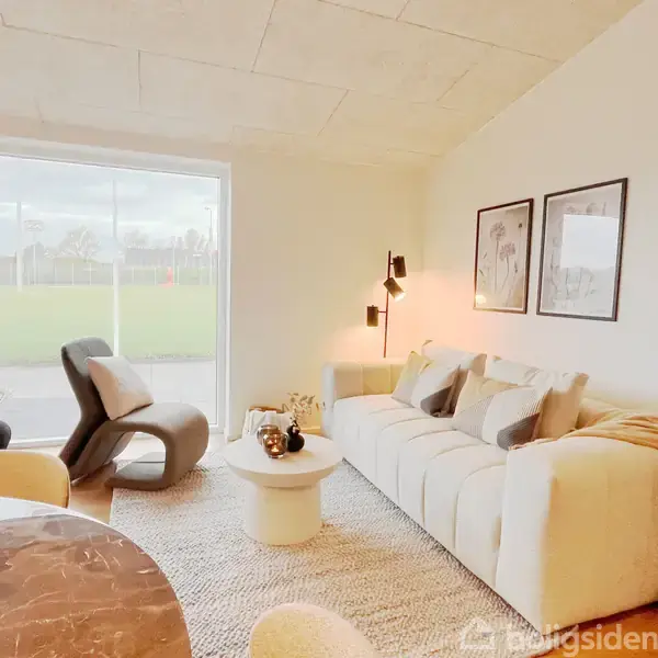 En lys stue med en beige sofa fyldt med puder, afslappende stol ved store vinduer, et rundt sofabord med en vase samt lamper og kunst på væggen.