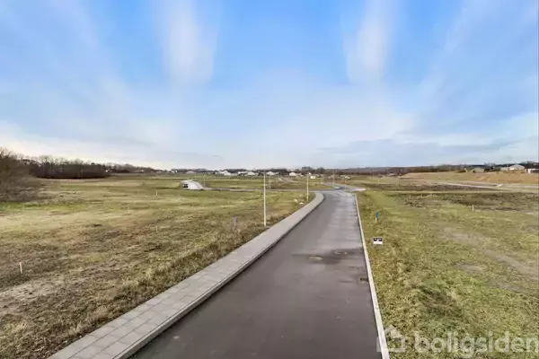 Asfaltvej løber gennem åbent landskab med græs og spredte buske. Langs vejen er der fortov til venstre, mens himlen er blå med spredte skyer.