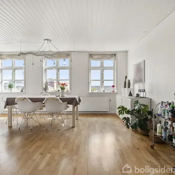 Spisestue med hvidmalet loft og vægge, stort bord og fire hvide stole, belyst af lysindfald fra flere vinduer. Til højre en skænk og plante, til venstre drikkeborde. Ingen tekst.