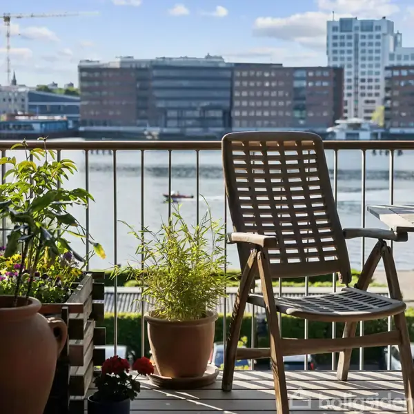 Stol står ved siden af blomsterkrukker på en altan. I baggrunden ses en kajakroer på en flod foran en by med høje bygninger.