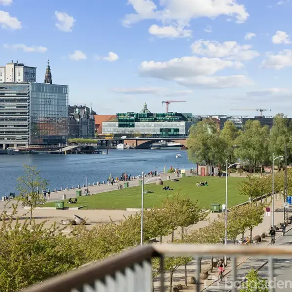 Flod strækker sig gennem bylandskab; mennesker nyder grønt område langs bredden. Moderne bygninger står på venstre side, mens en bro kan ses i baggrunden under en blå himmel.