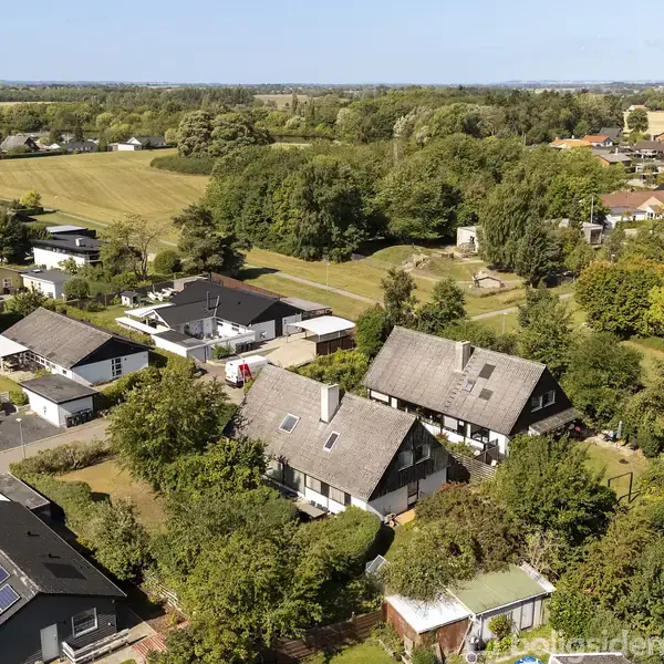 Et parcelhus med skråtag og solpaneler omgivet af frodige haver. Det ligger i et villakvarter med flere lignende huse, omgivet af marker og træer i en landlig byområde.