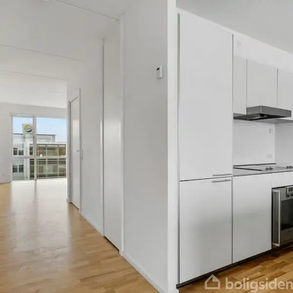 Et lille, moderne køkken med hvide skabe og indbygget ovn er placeret i en lys gang. Trægulve strækker sig ind i en stue med store vinduer mod en balkon.