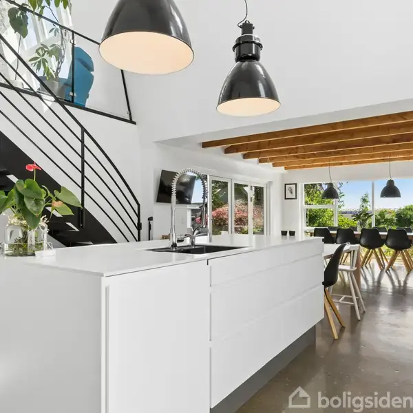 Et moderne køkken med en hvid ø og mørke pendellamper. En trappe fører til øverste etage. Store vinduer i spisestue med udsigt til en frodig have.