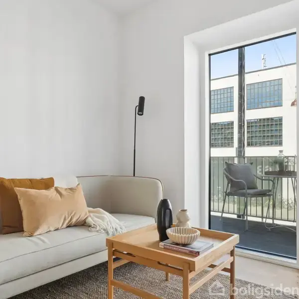 Sofa med puder står i et lyst rum med en gulvlampe ved siden af. Foran er et sofabord med dekoration. I baggrunden er franske døre til en altan med stole.