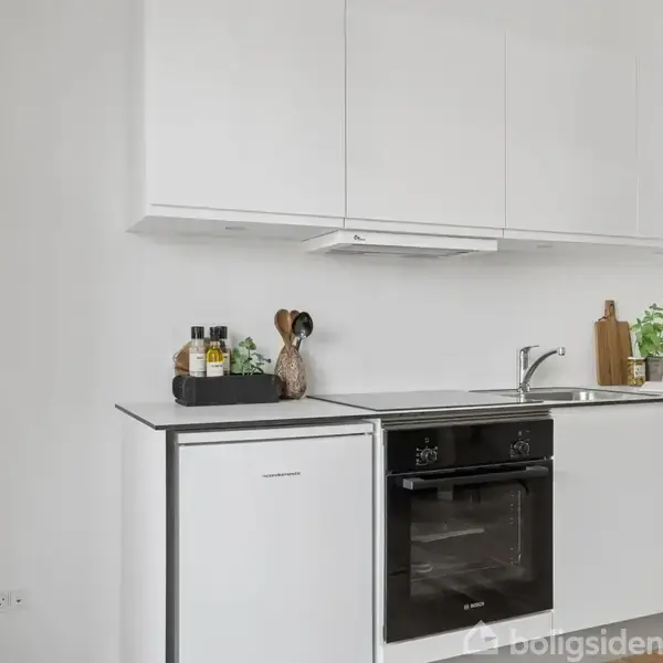 Et moderne køkken viser lyse skabe, en indbygget ovn og kogeplade. På bordpladen står krydderier og en træskærebræt. Rummet er lyst og minimalistisk indrettet.