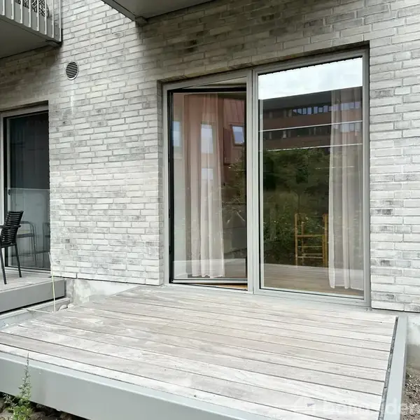 Glasdør med gardiner står i en muret murstensvæg. Ved siden af ses en sort stol. Træterrasse i forgrunden. Refleksioner viser et grønt udendørsområde.