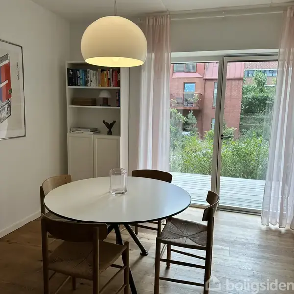 Et rundt bord med fire stole står i et lyst rum. En hylde med bøger står ved siden af, og store glasdøre åbner ud til en terrasse med udsigt over buske og bygninger.