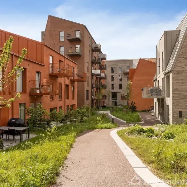 Række af moderne bygninger med røde og beige mursten står langs en sti i et grønt boligområde, omgivet af græs og små træer.