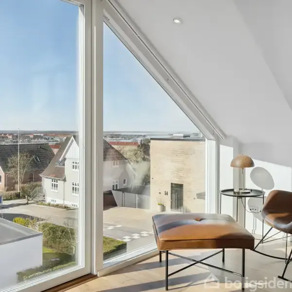 Et moderne rum med brune stole og bord placeret ved store, skråvinduer, der giver udsigt til et byområde med huse under en klar himmel.