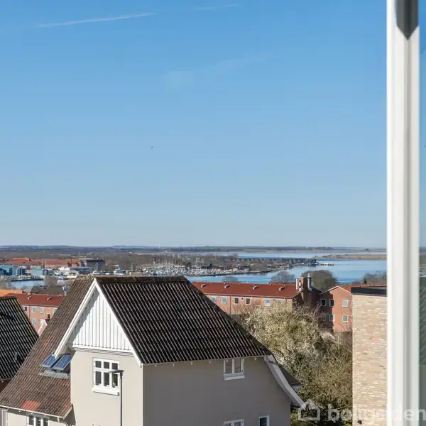 En udsigt fra et vindue viser hustage i en by med røde tage. I baggrunden ses en vandkant under en klar blå himmel.