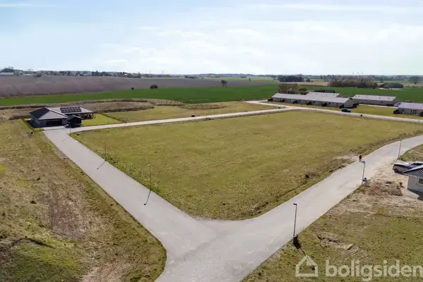 Et åbent græsareal er omkranset af asfalterede veje i et landligt område med spredte bygninger og marker i baggrunden under en blå himmel.