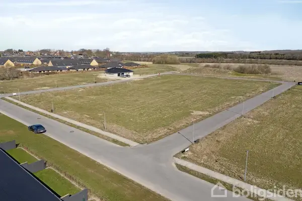 Åbent tomt græsareal ligger stille omkranset af veje i et beboelsesområde. I baggrunden ses rækkehuse og et skovområde, mens en blå bil er parkeret ved vejen.