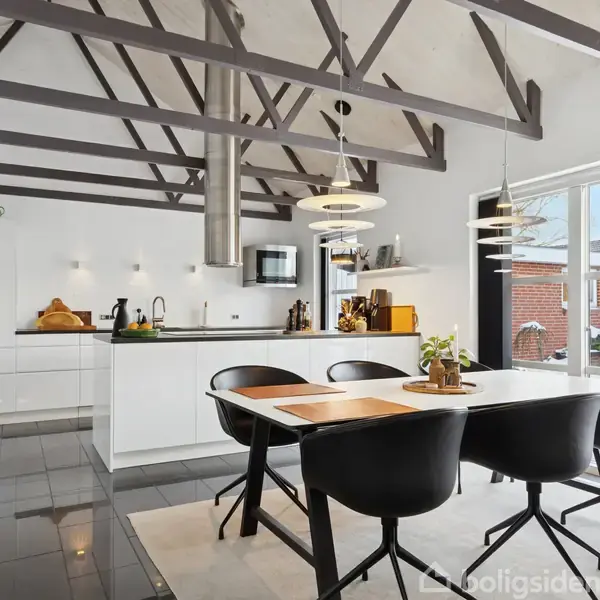 Køkken med sorte stole omkring et bord, omgivet af hvide skabe og apparater, belyst af hængende lamper. Store vinduer giver udsigt til en gårdhave. Maleri på væggen.