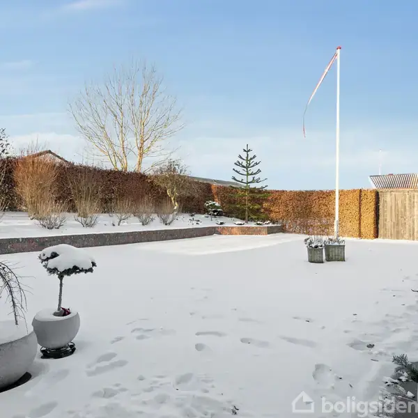 Sneklædt have med flagstang præsenterer rolige vinteromgivelser foran et hus med røde mursten. Bede med planter er delvist dækket af sne.