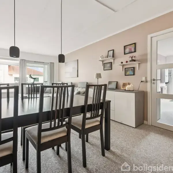 En spisebord med otte stole er centralt placeret i en lys spisestue. Til højre ses et skab med hylder og billeder, samt en dør ind til et tilstødende rum.